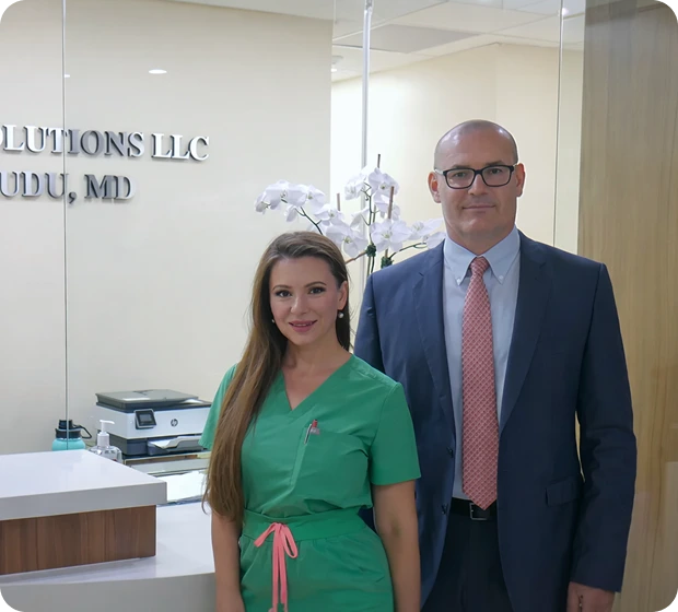 A woman in green scrubs stands beside a man in a suit and tie at an office reception desk. They both smile warmly, with orchids and office decor in the background.
