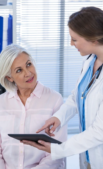 A female doctor in a white coat and blue scrubs shows a digital tablet to an older woman with white hair. They are engaged in a friendly discussion.